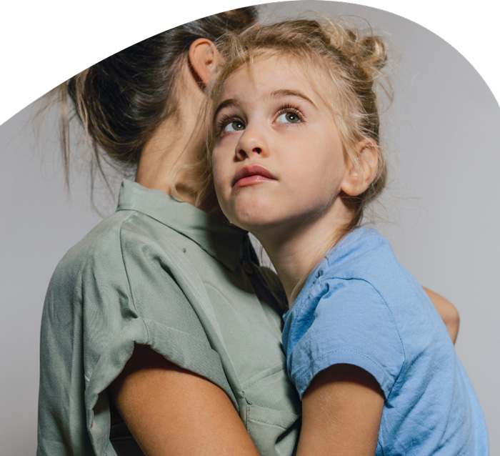 Ein kleines Mädchen umarmt eine erwachsene Person, deren Gesicht nicht zu sehen ist. Das Mädchen trägt ein blaues T-Shirt und schaut nach oben, mit einem Ausdruck der Nachdenklichkeit oder des Fragens. Der Hintergrund ist einheitlich und bietet einen starken Kontrast zu den Protagonisten des Bildes.